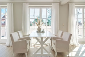 Un comedor con una mesa y sillas blancas, ventanas amplias con cortinas blancas, y vistas al exterior.