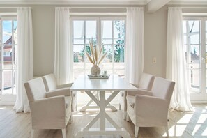 A dining area with a white table and chairs, large windows with white curtains, and a view of the outside.