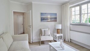 A modern, minimalist living room with a white sofa, a small side table with a lamp, a painting of a beach scene, and a large window with curtains. - Welcome to TECHTS - TECHTs Strandhaus (Timmendorfer Strand)