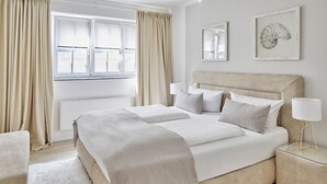 A hotel room with a large bed, beige curtains, a bedside table with a lamp, and two framed pictures on the wall. - Welcome to TECHTS - TECHTs Strandhaus (Timmendorfer Strand)