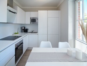 A modern kitchen with white cabinets, a built-in microwave, a coffee maker, and a sink. There are two white chairs and a table with a grey placemat. - A Love for the Coast – The Baltic Sea Up Close - TECHT's Beach House (Timmendorfer Strand)