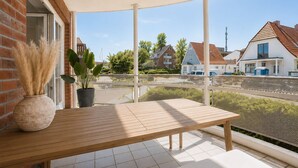 A balcony with a wooden table and chairs, potted plants, and a view of a residential street. - Welcome to TECHTS - TECHTs Strandhaus (Timmendorfer Strand)