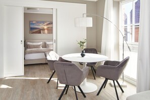 Una habitación de hotel moderna con una mesa de comedor redonda, cuatro sillas, un sofá junto a la cama, un ventanal con cortinas y un cuadro en la pared.