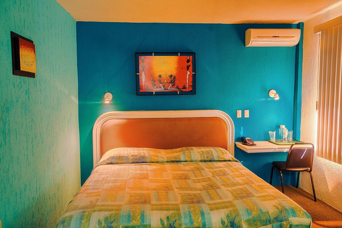 Ein Zimmer mit einem Bett, einem Schreibtisch und einem Stuhl. An der Wand hängt ein Bild und über dem Schreibtisch befindet sich eine Klimaanlage.
