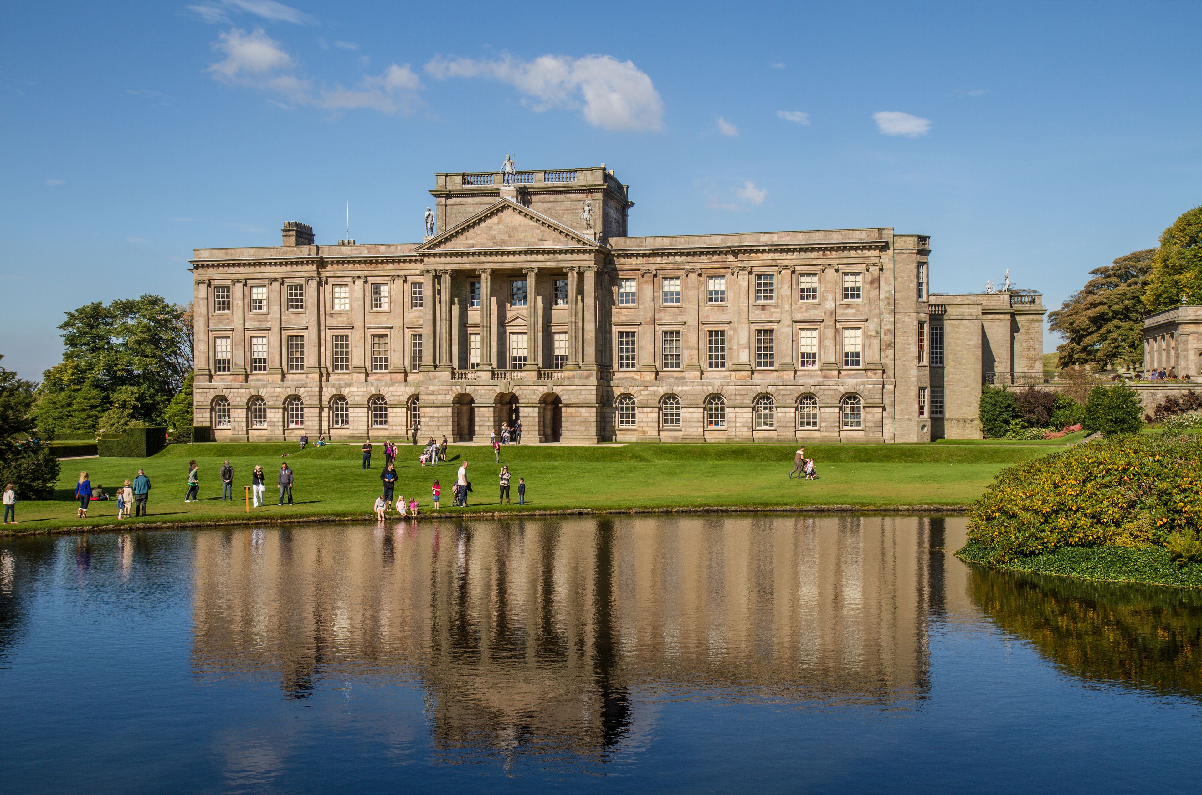 A large, historic building with a symmetrical facade, a central entrance with columns, and a reflection in the water in front.