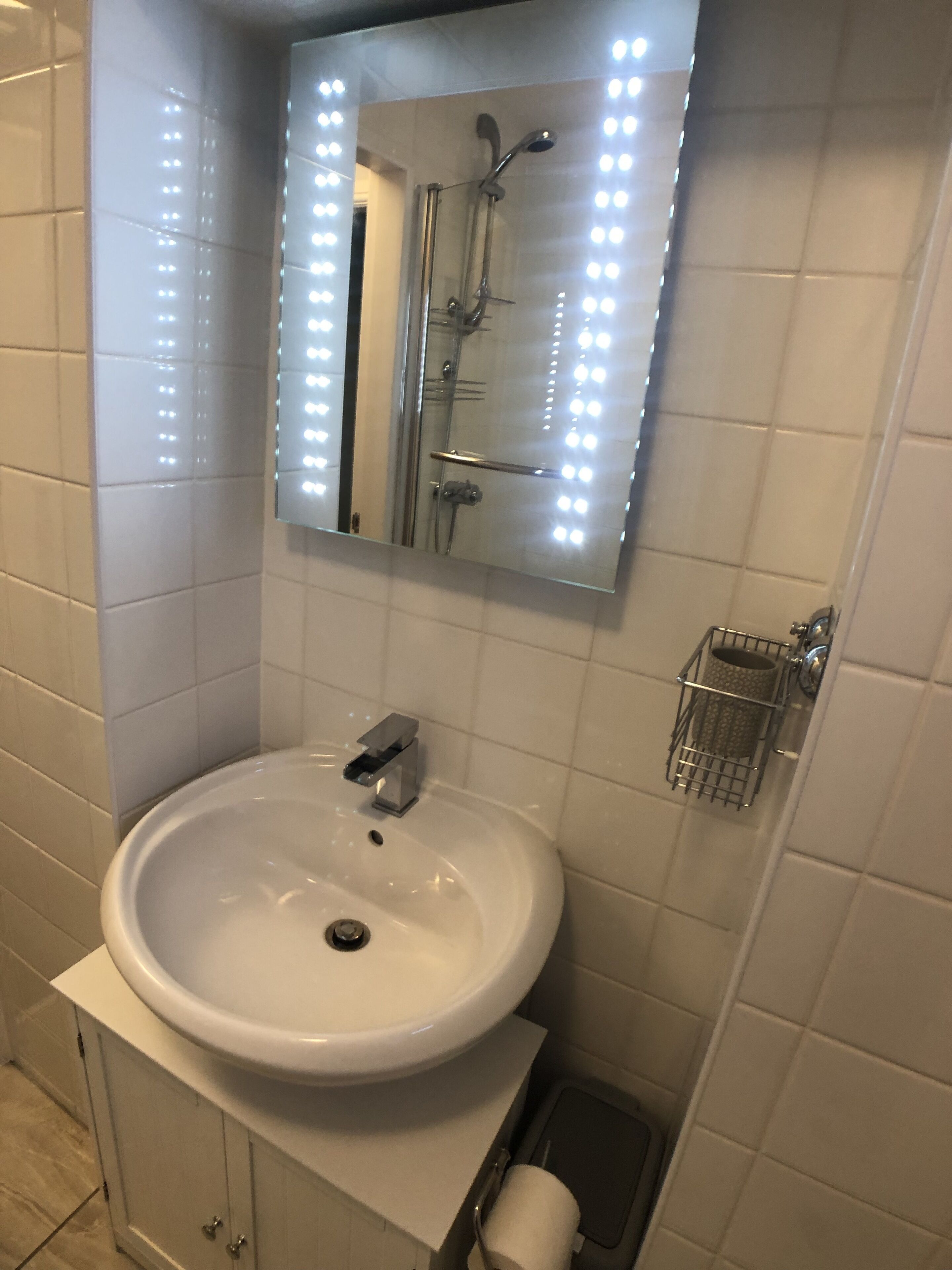 A bathroom with a white sink, a mirror with lights, a shower, and a toilet paper holder.