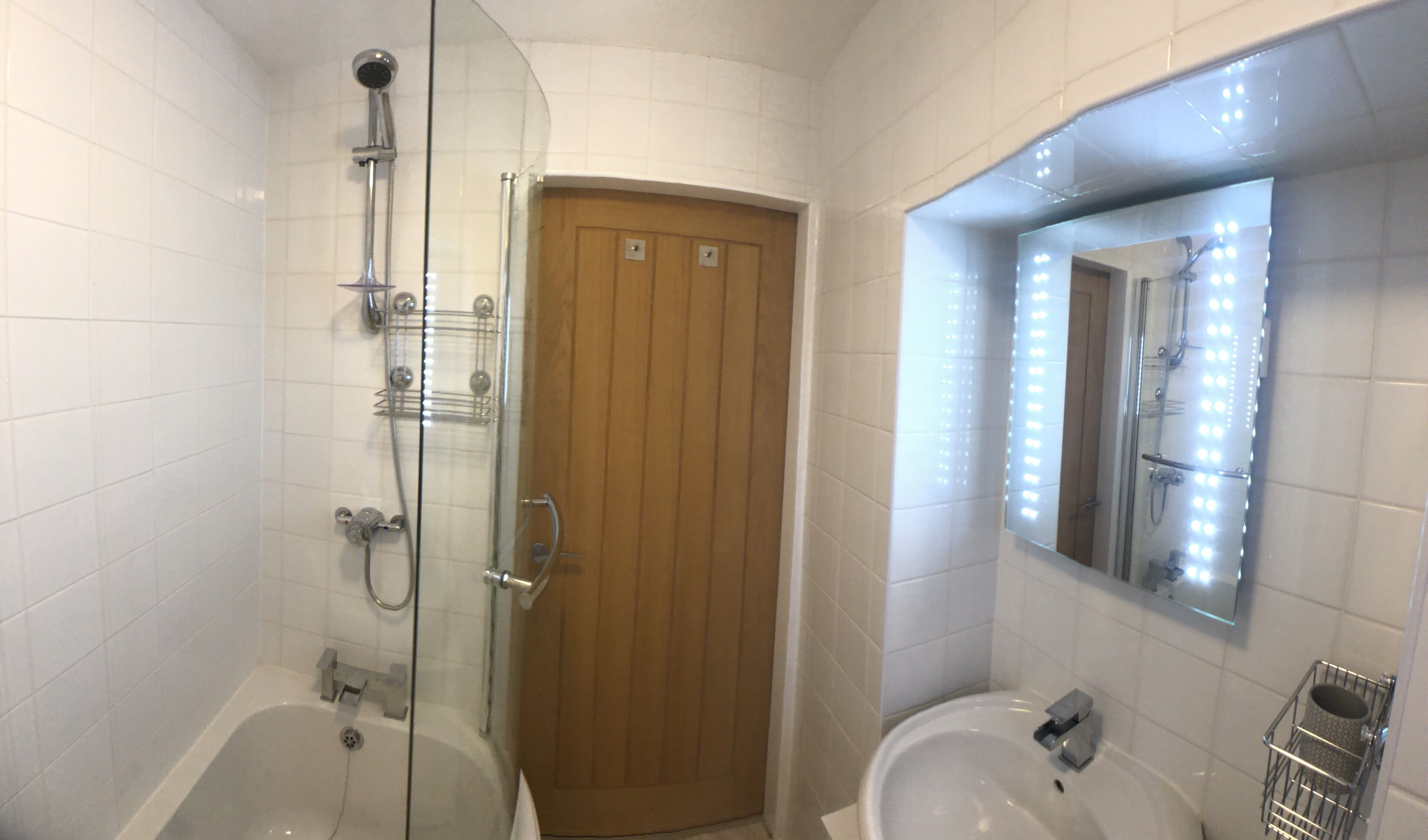 A bathroom with a glass-enclosed shower, a wooden door, a sink, and a mirror with lights.