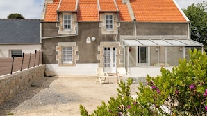 Une maison à deux étages avec un toit recouvert de tuiles rouges, un petit porche avec des chaises et une allée gravillonnée.