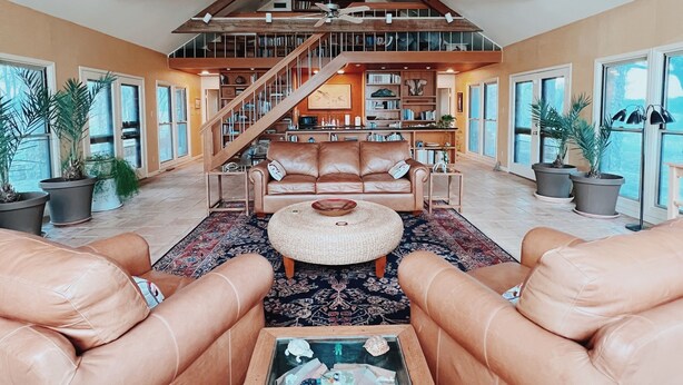 A spacious living room with a high ceiling, wooden beams, a leather sofa, a coffee table with decorative items, and a staircase leading to an upper level. - M'walimu 4 BR w/ Pool, Farm Stay- Flower Farm, Goats, Pigs, Disc Golf! (Glen Rose)