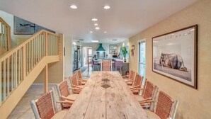 A spacious dining area with a long wooden table and wicker chairs, a staircase with wooden railings, and a kitchen in the background. - M'walimu 4 BR w/ Pool, Farm Stay- Flower Farm, Goats, Pigs, Disc Golf! (Glen Rose)