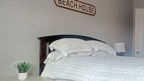 A bedroom with a bed, a nightstand, a potted plant, a wall sign reading "BEACH HOUSE", and a door. - Pet-friendly guest suite. Private fenced yard. 2 min walk to beach.  (Carolina Beach)