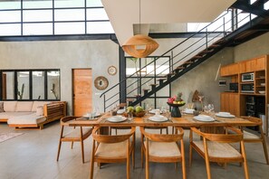 Un comedor espacioso con una larga mesa de madera dispuesta para seis personas, flanqueada por sillas de madera. Una escalera con barandilla negra conduce a un nivel superior.