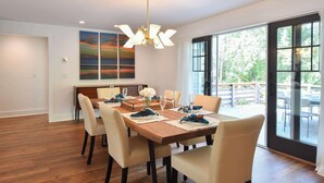 A dining area with a wooden table set for six, surrounded by beige chairs. A modern chandelier hangs from the ceiling, and a large window offers a view of the outdoors. - Luxurious, Modern Walk to Water! (Orleans)