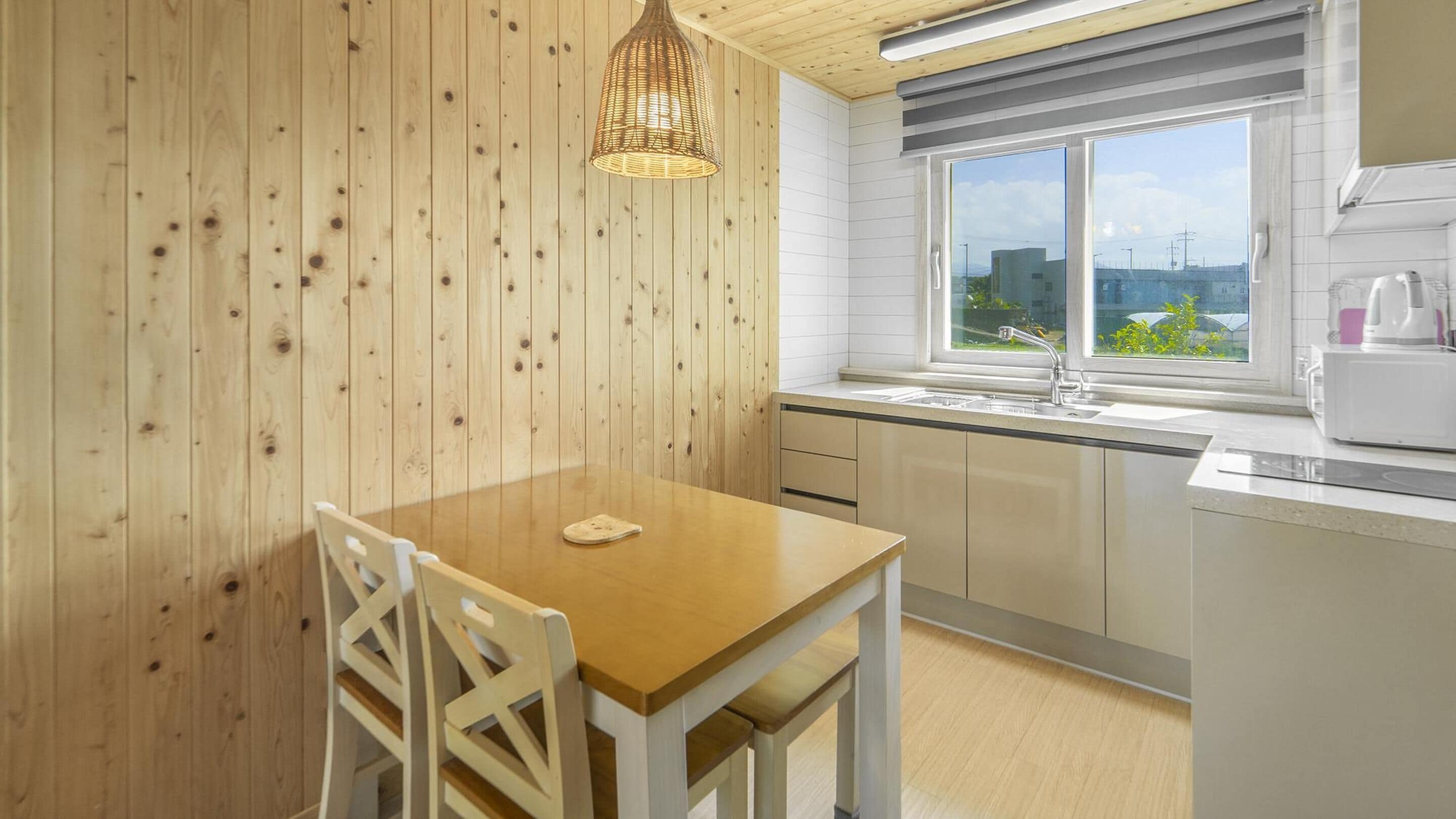 A modern kitchen with wooden walls, a wooden dining table, white chairs, a window with a view, and a white microwave.