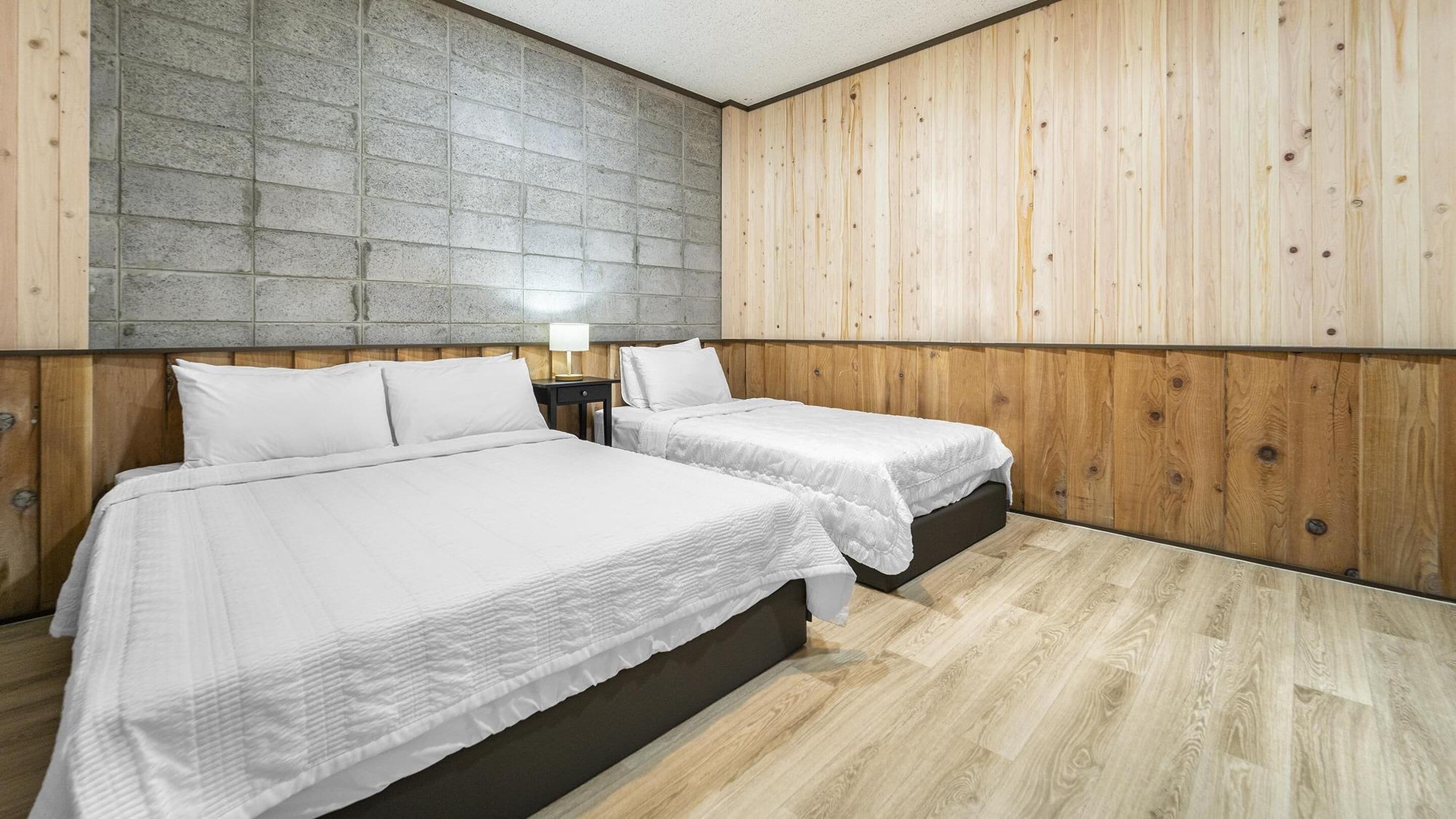 A room with two beds, a stone wall, wooden paneling, and a nightstand with a lamp.