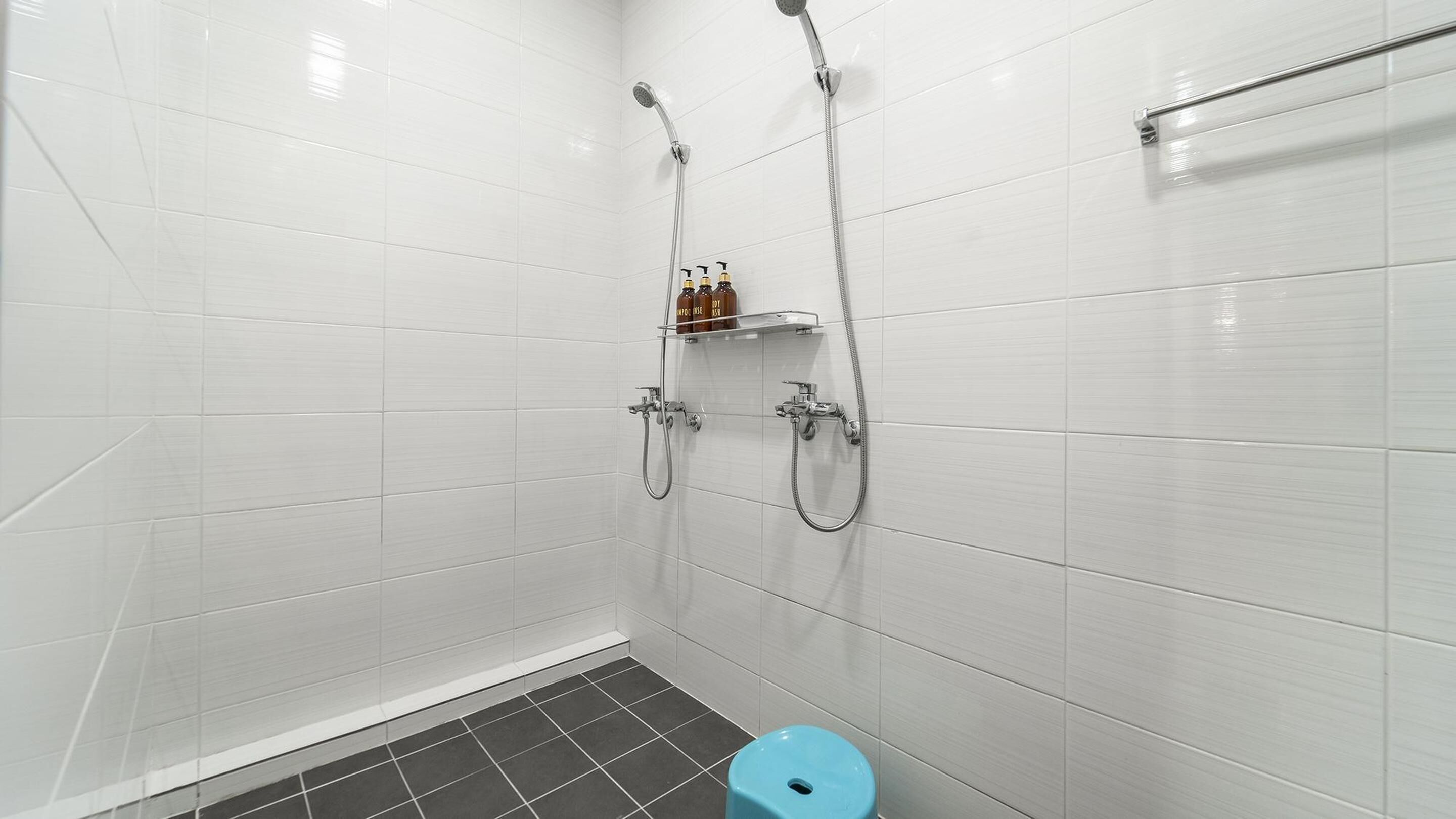 A small, modern bathroom with white tiled walls and a black tiled floor. There are two showerheads and a toilet.