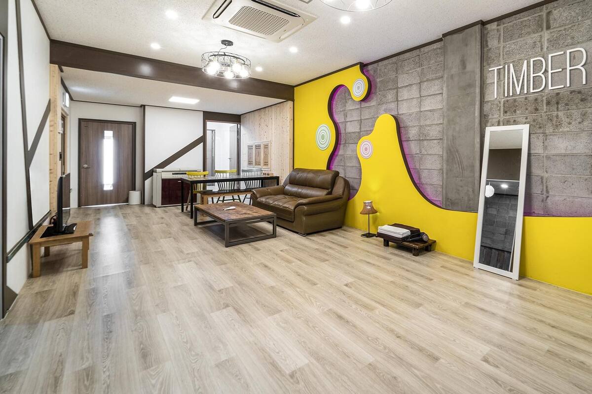 Un vaste espace de vie comprenant un canapé en cuir marron, une table basse et une table à manger avec des chaises. Un grand mur décoré d’une fresque jaune et violette sur lequel on peut lire le mot « TIMBER » est visible.