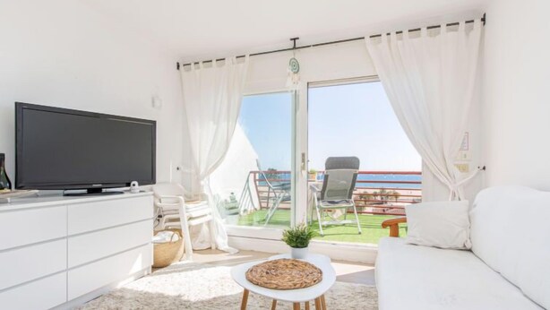Un salon moderne doté d’un téléviseur à écran plat, d’un canapé blanc et d’un balcon offrant une vue sur la mer.