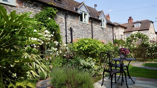 Una casa de piedra con un patio jardín, una mesa con sillas y una variedad de plantas y flores.