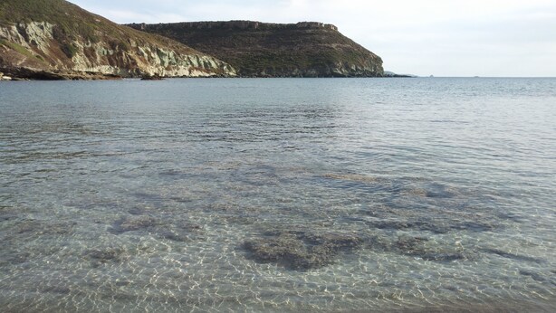 Ein Küstenstrand mit klarem, flachem Wasser, durch das der sandige Meeresboden sichtbar ist, ein Felsvorsprung im Hintergrund und eine weite Ansicht auf das Meer.