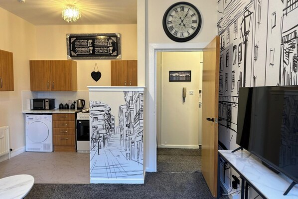 Een kleine keuken met houten kastjes, een magnetron, een koelkast en een klok aan de muur.