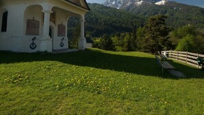 Une église surmontée d’une croix, un banc et une clôture, le tout situé dans un espace verdoyant avec une montagne en arrière-plan.
