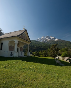 A church with a cross on the roof, a bench, and a fence in a grassy area with a mountain in the background. - Vacation apartment Serlesblick with direct access to the ski slope, WLAN and terrace (Mieders)