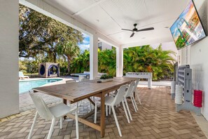 An outdoor patio with a wooden table and white chairs, a pool, and a TV mounted on the wall.