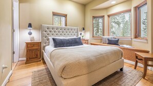 A bedroom with a large bed, two bedside tables with lamps, a window seat, and a view of trees. - Sleeps 20 | Hot Tub + Game Room & Theater | Steps to Ski Shuttle | River Rock (Breckenridge)