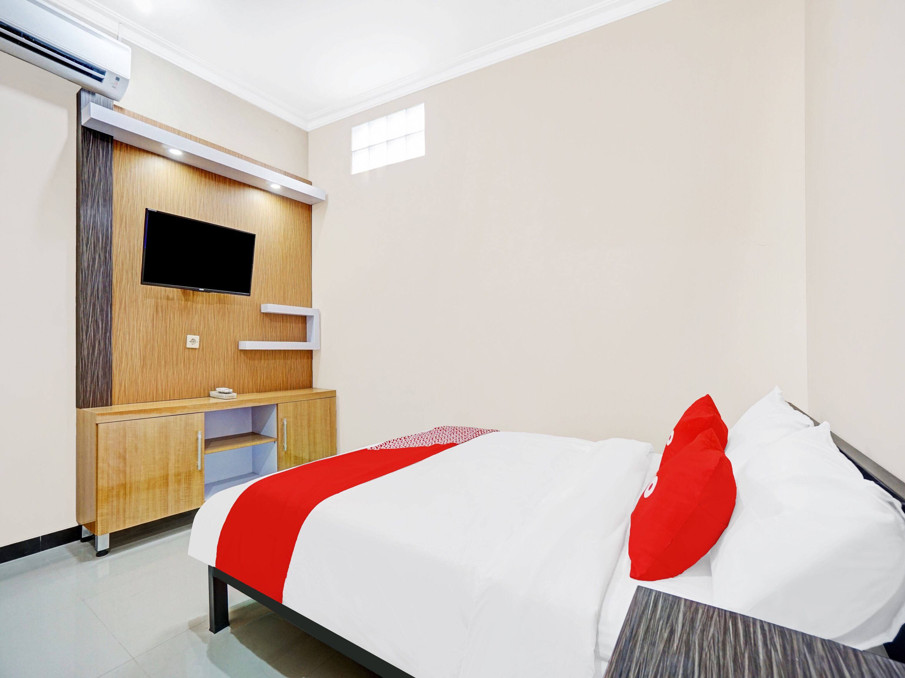 A hotel room with a bed, a TV mounted on a wooden panel, a small desk, and an air conditioning unit.
