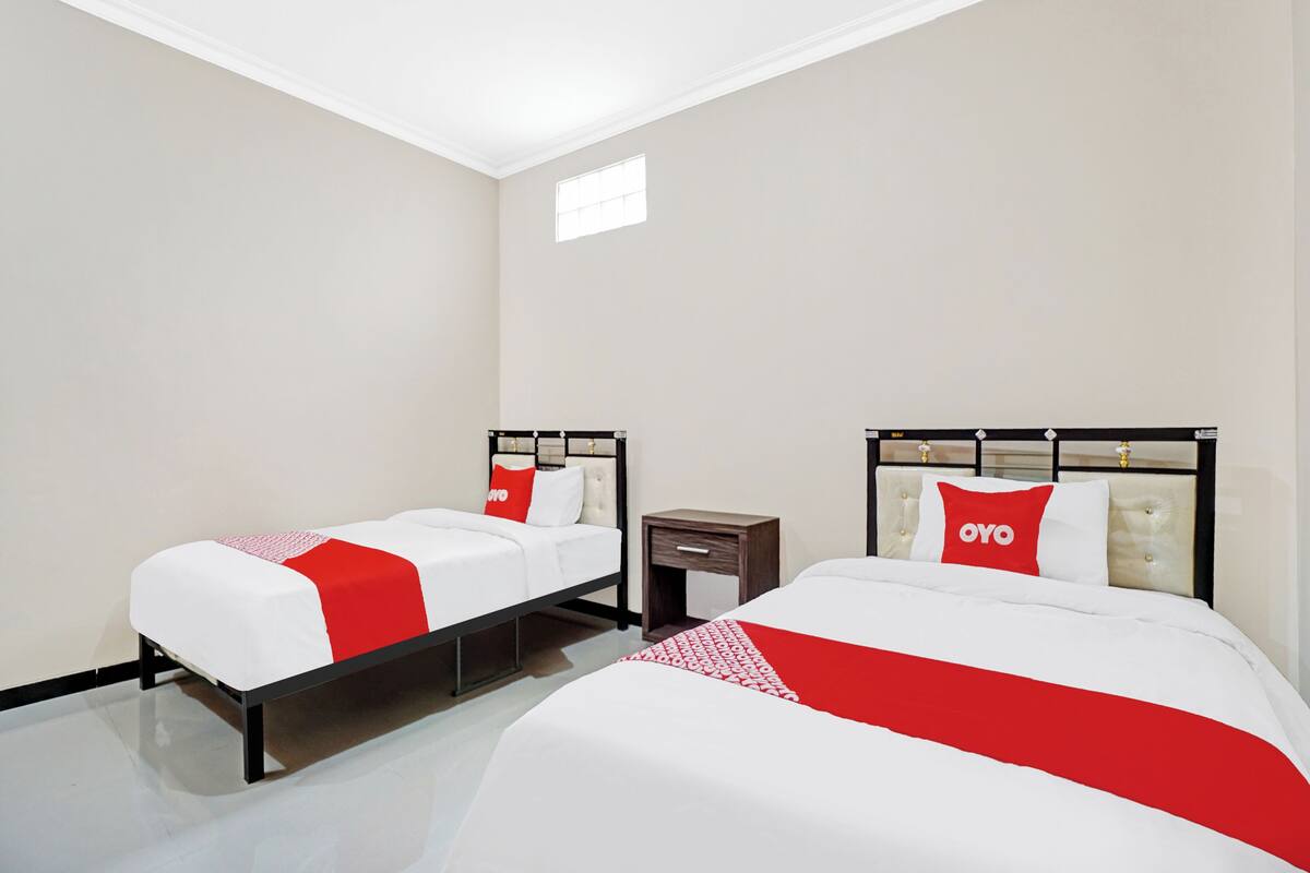 Ein Hotelzimmer mit zwei Betten, jeweils mit einer roten und weißen Bettdecke und passenden Kissen. Das Zimmer hat ein modernes Design mit weißer Decke und Wänden sowie einem kleinen Fenster, das natürliches Licht hereinlässt.