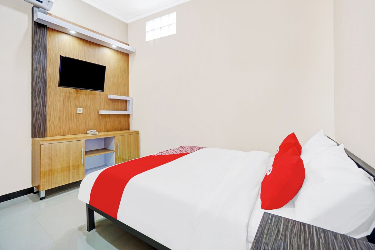A hotel room with a bed, a TV mounted on a wooden panel, a small desk, and an air conditioning unit.
