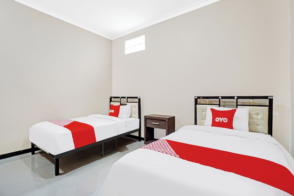 A hotel room with two beds, each with a red and white bedspread and matching pillows. The room has a modern design with a white ceiling and walls, and a small window providing natural light.
