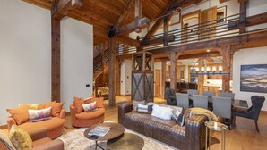 A spacious living area with a high ceiling, wooden beams, and a cozy seating arrangement including sofas and armchairs. A staircase leads to an upper level, and a dining area is visible in the background. - Private Ski in/out Lodge! Hot Tub, 2 King En Suites, Stunning Views (Mountain Village)