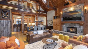 A spacious living area with a fireplace, wooden beams, a dining area, and a kitchen visible through an open archway. - Private Ski in/out Lodge! Hot Tub, 2 King En Suites, Stunning Views (Mountain Village)