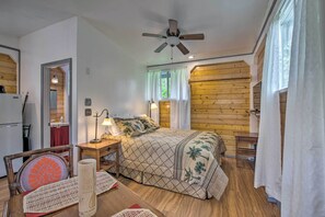 Ein Schlafzimmer mit einem Bett, Holzwänden, einem Deckenventilator, einem Nachttisch mit Lampe und Blick auf die Küche.
