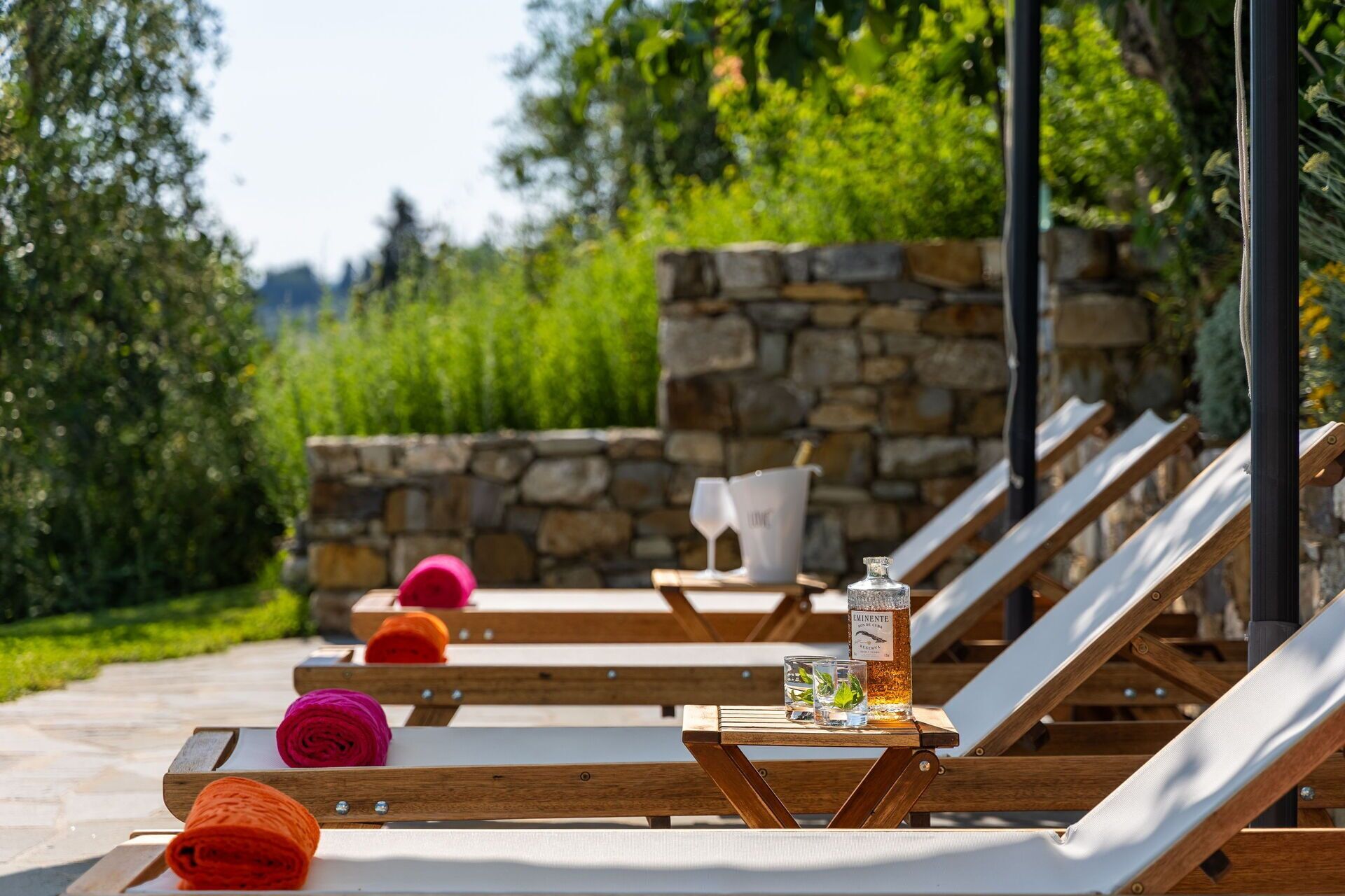 Outdoor lounge chairs with cushions, a table with a bottle and glasses, and a stone wall in the background.