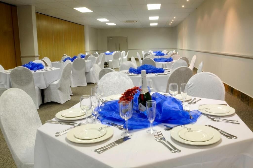 Een feestzaal met ronde tafels gedekt voor een diner, elk met witte tafelkleden, bestek en blauwe stoelhoezen.