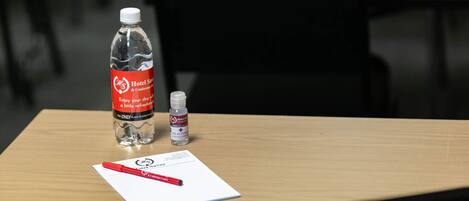 Una habitación de hotel con una mesa de madera, una botella de agua, un pequeño frasco de gel hidroalcohólico y un bolígrafo sobre un papel.