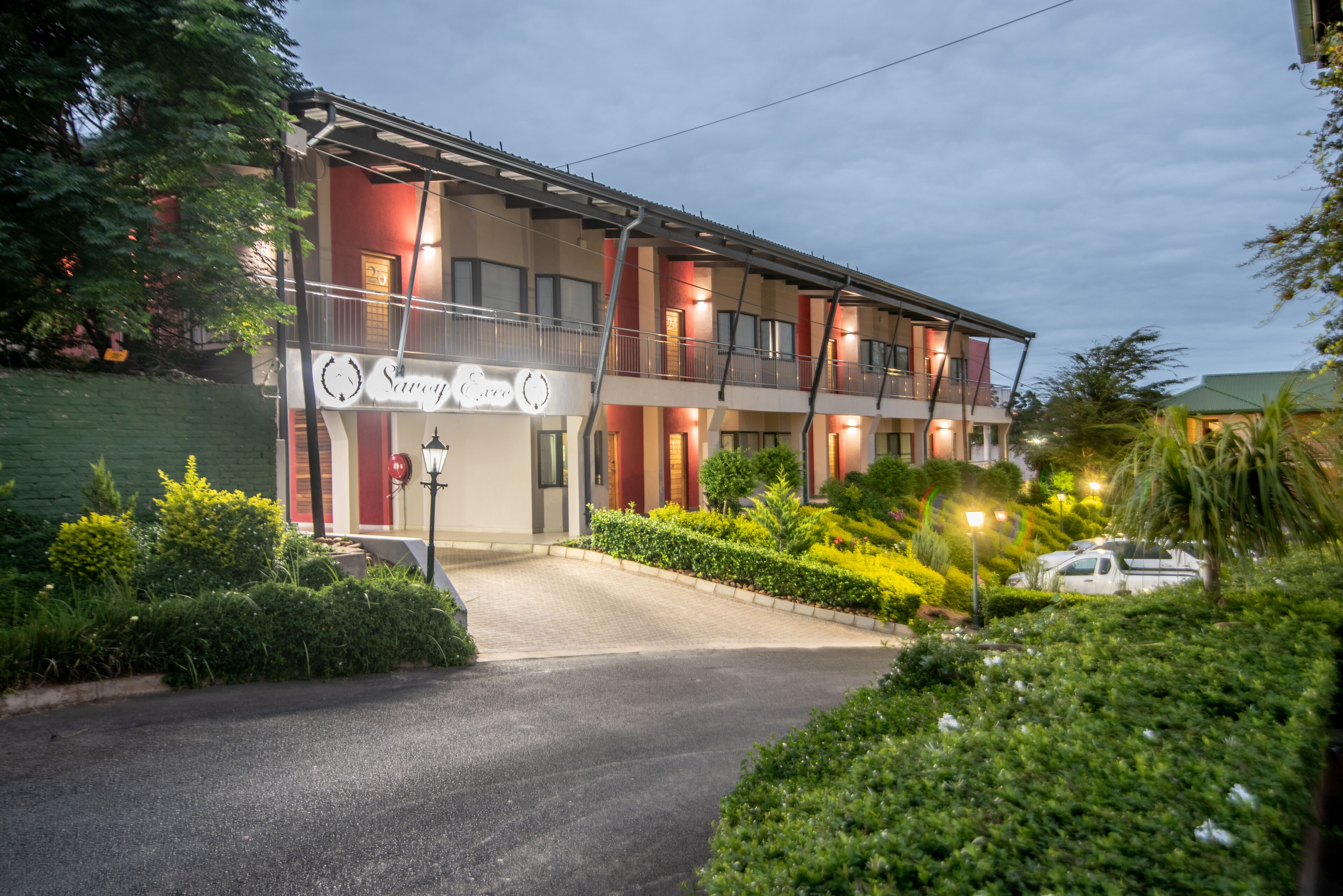 A multi-story hotel with a balcony on the second floor, a well-maintained garden, and a clear driveway.