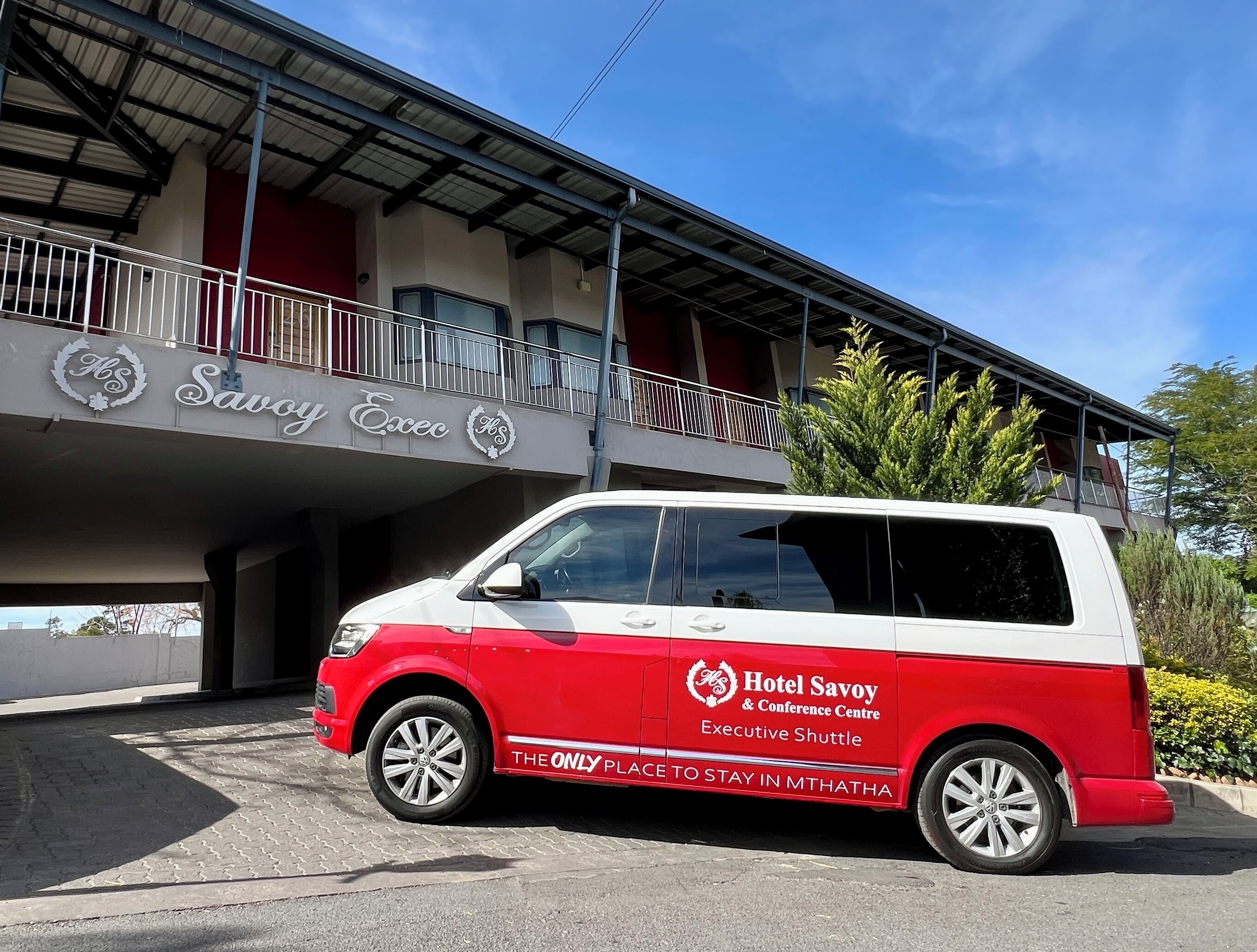 A red and white shuttle bus parked in front of a building with a sign that reads 'Hotel Savoy & Conference Centre Executive Shuttle.'