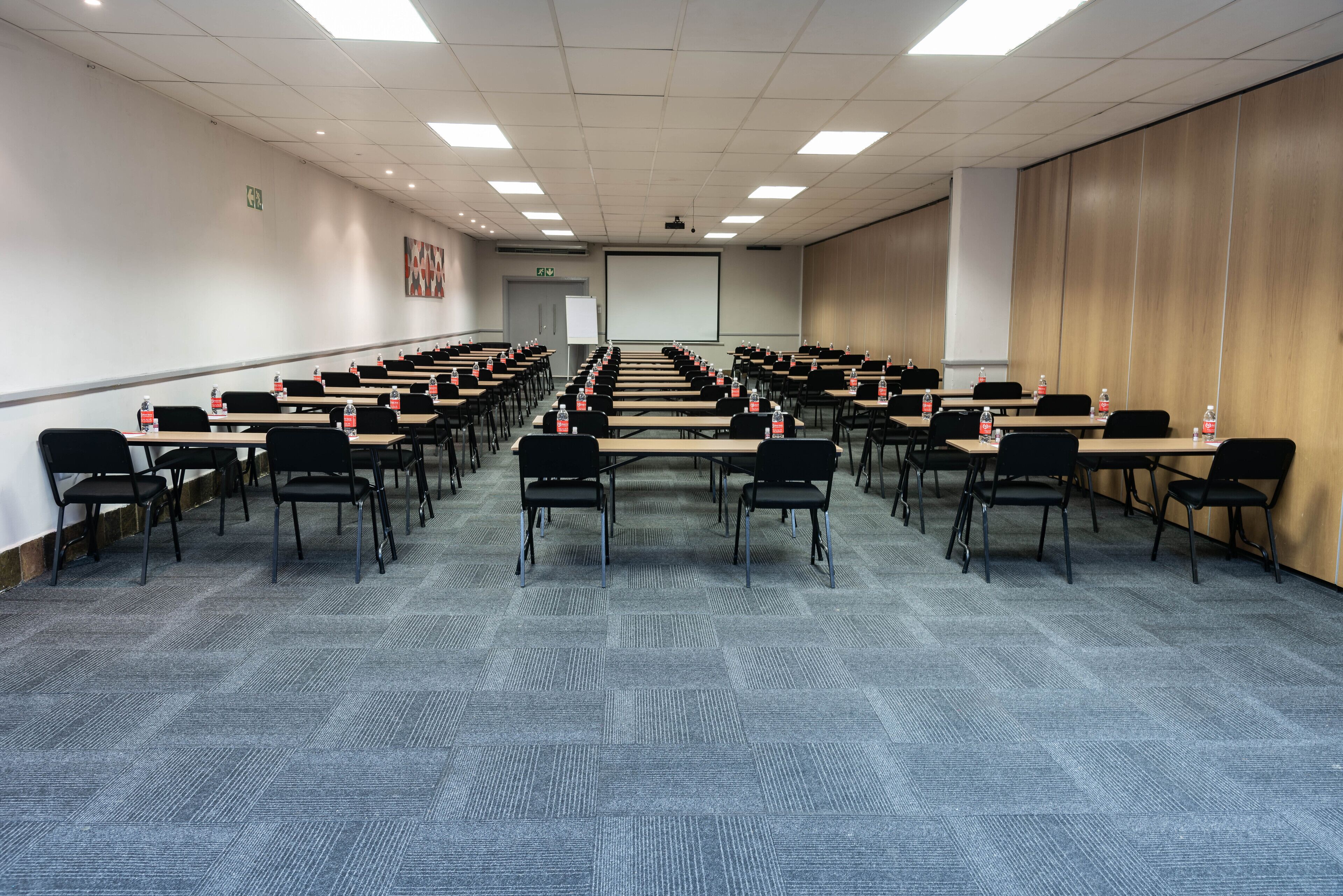 Une salle de conférence avec des rangées de tables et de chaises, équipée de bouteilles d’eau, d’un écran de projection et d’un tableau blanc.