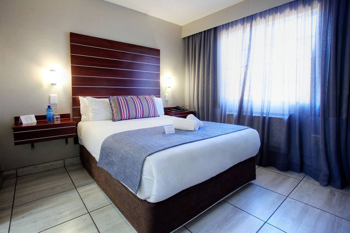 Hotelkamer met een groot bed, houten hoofdeinde, twee nachtkastjes met lampen, een bijzettafeltje en een raam met gordijnen.