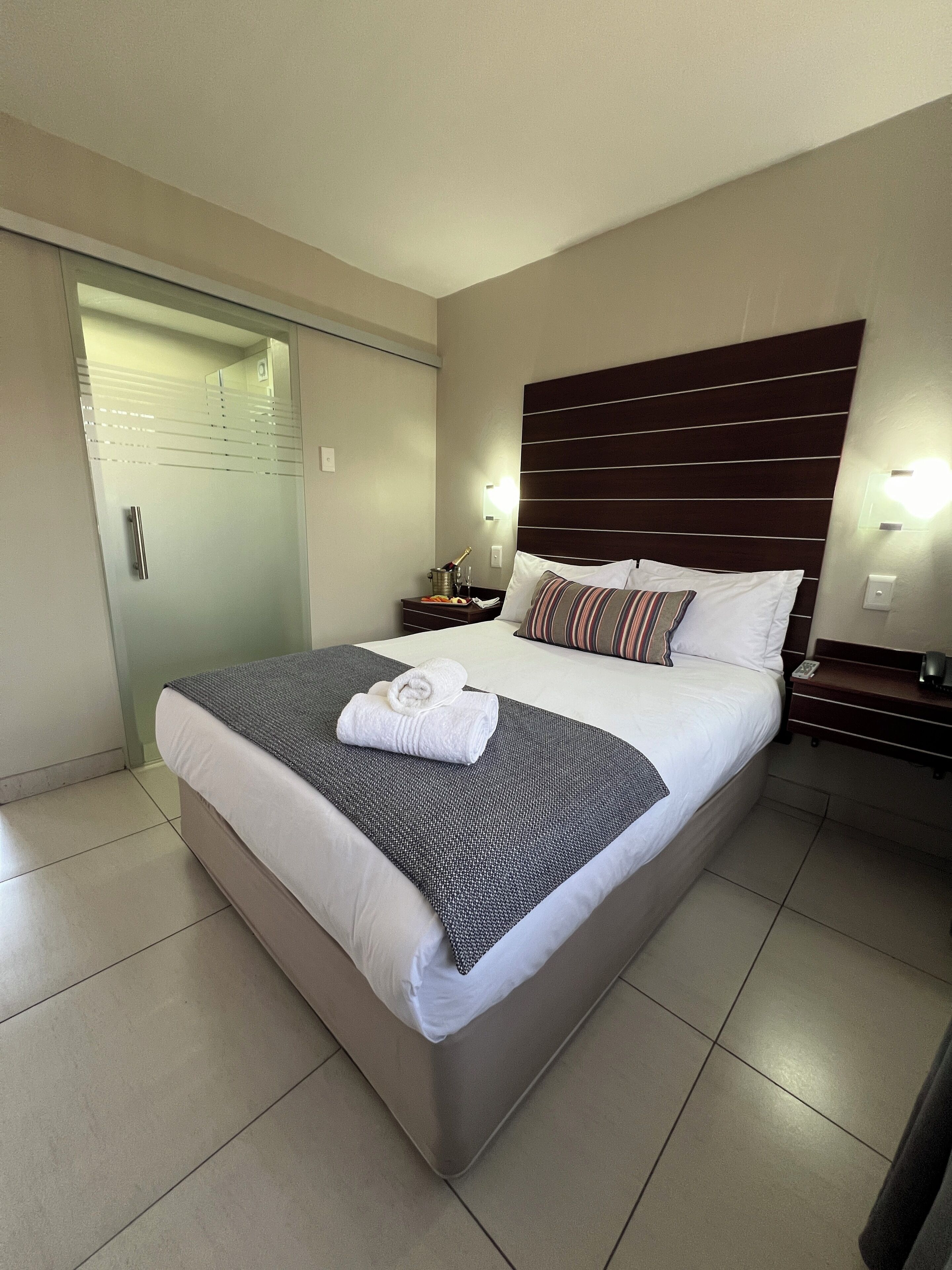 A neatly made bed with a striped pillow, a folded towel, and a bedside table with a lamp and a bottle.