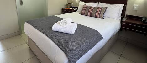 Una cama bien tendida, con un cojín a rayas, una toalla doblada y una mesita de noche con una lámpara y una botella.