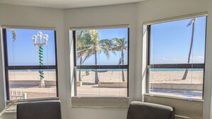 Une pièce dotée de trois grandes fenêtres donnant sur une plage bordée de palmiers et d’une étendue de sable. On y trouve deux fauteuils rembourrés et une petite table sur laquelle est posé un objet décoratif.
