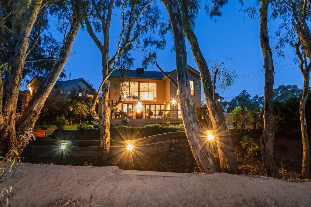 A house with a large window, a porch, and a well-lit front yard at dusk.