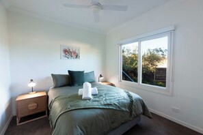 A bedroom with a bed, two bedside tables, a ceiling fan, a window with a view, and a wall-mounted artwork.