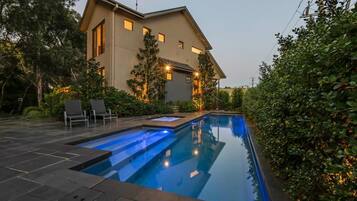 A modern house with a swimming pool, surrounded by greenery and two lounge chairs.