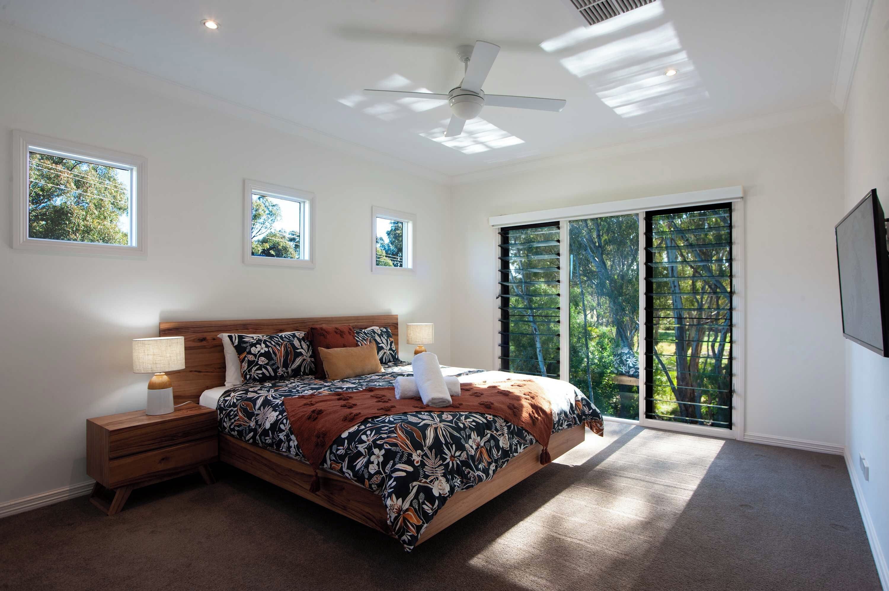 A bedroom with a large bed, two bedside tables, a ceiling fan, and a view of trees through the window.